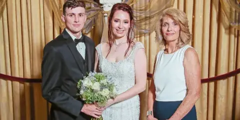 A bride and groom standing with a female wedding officiant in front of a gold curtain backdrop.