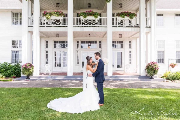 A bride and groom standing on a lawn in front of a large white historic venue building with a columned porch.
