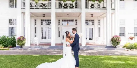 A bride and groom standing on a lawn in front of a large white historic venue building with a columned porch.