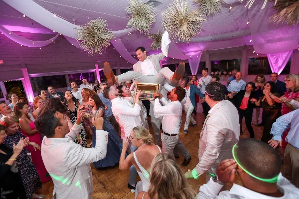 A crowded wedding reception dance floor with guests cheering as a groom is tossed on a chair.
