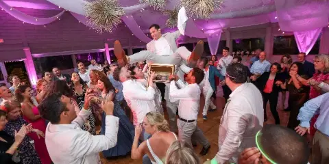 A crowded wedding reception dance floor with guests cheering as a groom is tossed on a chair.