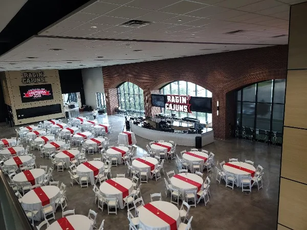 An indoor event venue set up with many round tables covered in white tablecloths and red runners, surrounded by white chairs.