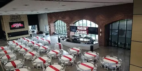 An indoor event venue set up with many round tables covered in white tablecloths and red runners, surrounded by white chairs.