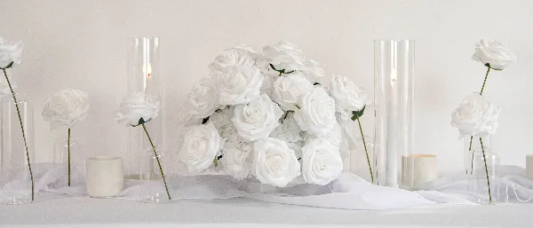 A wedding table setting featuring a large arrangement of white roses, tall glass candle holders, and individual white roses in glass vases on a white tablecloth.