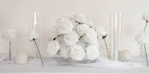 A wedding table setting featuring a large arrangement of white roses, tall glass candle holders, and individual white roses in glass vases on a white tablecloth.