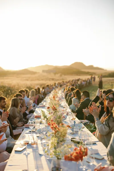 A long outdoor wedding reception table with floral centerpieces and guests seated on both sides under a sunset sky.