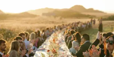 A long outdoor wedding reception table with floral centerpieces and guests seated on both sides under a sunset sky.