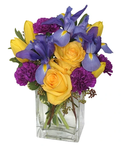 A floral arrangement featuring yellow roses, purple irises, and purple carnations in a square glass vase.