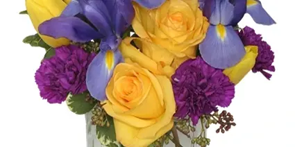 A floral arrangement featuring yellow roses, purple irises, and purple carnations in a square glass vase.