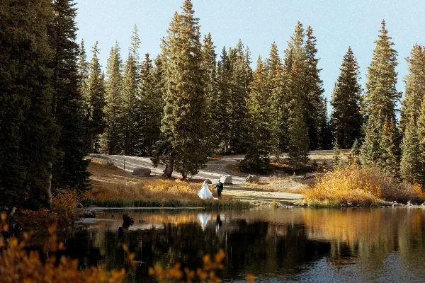 A bride and groom standing by a calm lake surrounded by tall pine trees and autumn foliage.