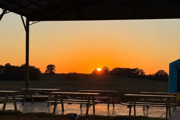A covered outdoor pavilion with wooden picnic tables set up in a rural field at sunset.