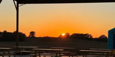 A covered outdoor pavilion with wooden picnic tables set up in a rural field at sunset.