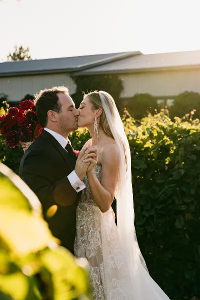 A bride and groom kissing outdoors in golden hour light, with the bride holding a bouquet of dark red flowers.
