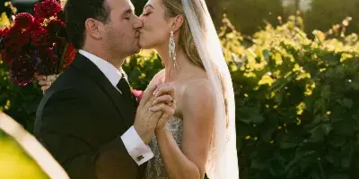 A bride and groom kissing outdoors in golden hour light, with the bride holding a bouquet of dark red flowers.