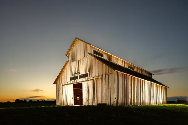 A large wooden barn with a steep roof and open doorway illuminated at dusk in a rural field.