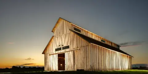 A large wooden barn with a steep roof and open doorway illuminated at dusk in a rural field.