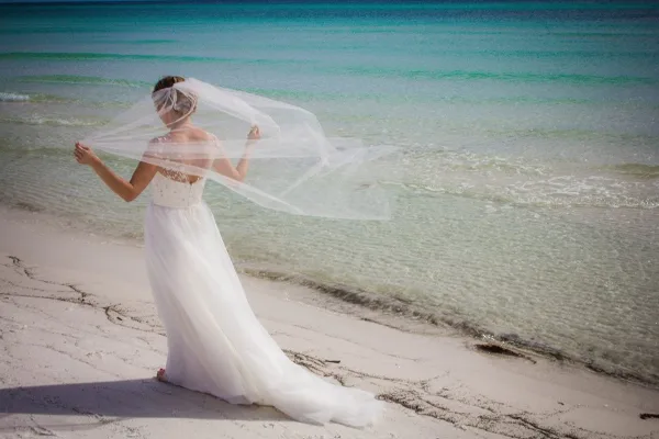 A bride in a white wedding dress and veil standing on a sandy beach with turquoise ocean water in the background.