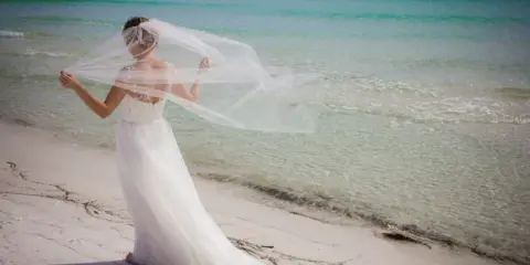 A bride in a white wedding dress and veil standing on a sandy beach with turquoise ocean water in the background.