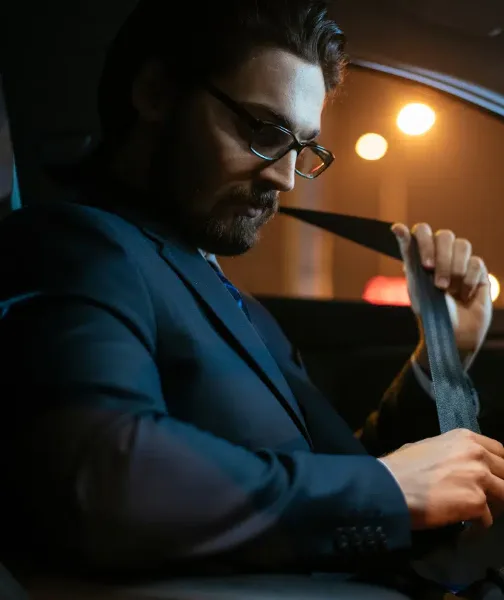 A man in a suit and glasses fastening a seatbelt inside a car.