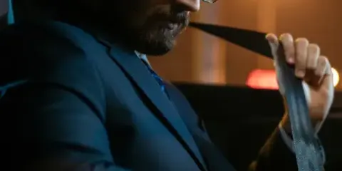 A man in a suit and glasses fastening a seatbelt inside a car.