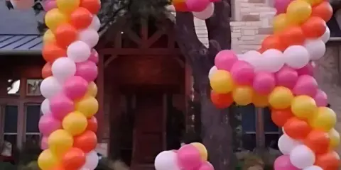 Large outdoor balloon letters in pink, orange, yellow, and white arranged on a lawn in front of a house.