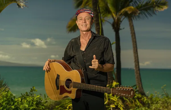 A smiling man in a black shirt and bandana holding an acoustic guitar outdoors with palm trees and the ocean in the background.