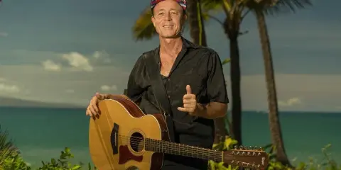 A smiling man in a black shirt and bandana holding an acoustic guitar outdoors with palm trees and the ocean in the background.