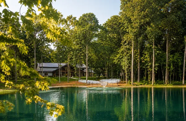 A modern venue building surrounded by a forest with a lakeside ceremony area featuring white chairs.