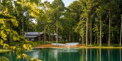 A modern venue building surrounded by a forest with a lakeside ceremony area featuring white chairs.