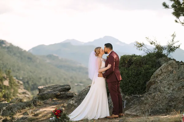 A bride and groom sharing a kiss on a rocky mountain overlook with a scenic mountain range in the background.