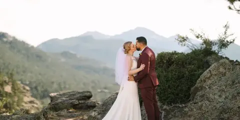 A bride and groom sharing a kiss on a rocky mountain overlook with a scenic mountain range in the background.