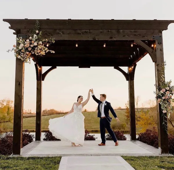 A bride and groom dancing under a wooden pergola at an outdoor wedding ceremony site overlooking a pond.