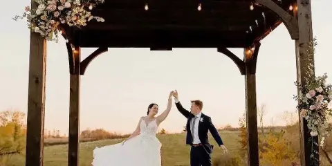 A bride and groom dancing under a wooden pergola at an outdoor wedding ceremony site overlooking a pond.