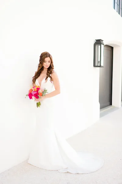 Bride in a white gown holding a bright pink and orange floral bouquet against a white wall