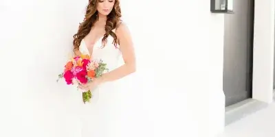 Bride in a white gown holding a bright pink and orange floral bouquet against a white wall
