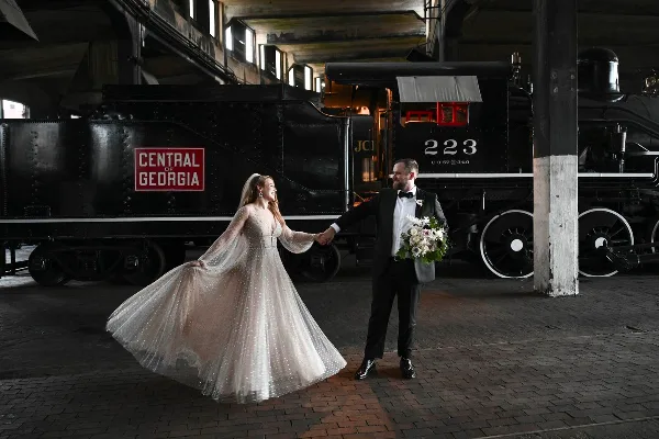 A bride and groom holding hands and smiling in front of a black steam locomotive at the Georgia Railroad Museum.