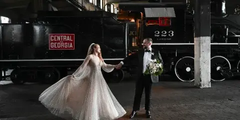 A bride and groom holding hands and smiling in front of a black steam locomotive at the Georgia Railroad Museum.