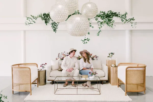 A couple sitting on a white sofa in a styled event space with rattan chairs, a glass coffee table, and hanging paper lanterns.