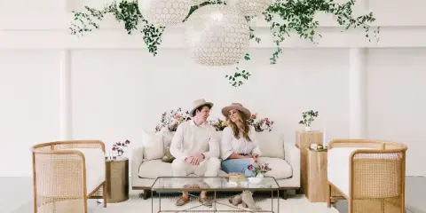 A couple sitting on a white sofa in a styled event space with rattan chairs, a glass coffee table, and hanging paper lanterns.