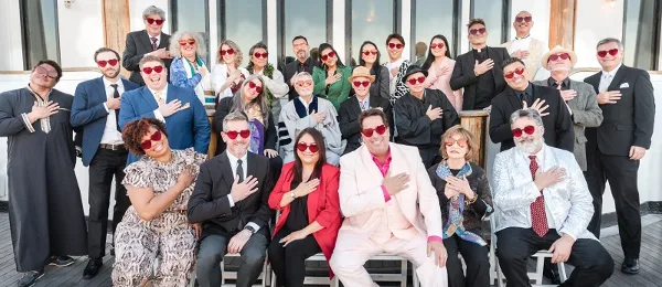 A large group of people in formal and themed attire, including suits, dresses, and a priest's robe, posing together outdoors.