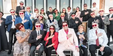 A large group of people in formal and themed attire, including suits, dresses, and a priest's robe, posing together outdoors.