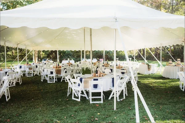 A large white event tent set up on a grassy lawn containing multiple round tables with white tablecloths and white folding chairs.