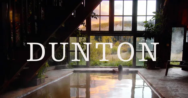 Interior view of a rustic resort pool with a person soaking, looking out large windows at a mountain landscape at sunset.