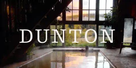 Interior view of a rustic resort pool with a person soaking, looking out large windows at a mountain landscape at sunset.