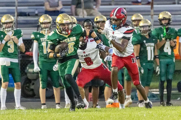 A high school football player in a green Titans uniform carrying the ball while being pursued by a player in a red East uniform.