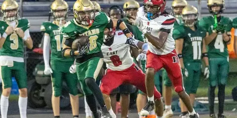 A high school football player in a green Titans uniform carrying the ball while being pursued by a player in a red East uniform.