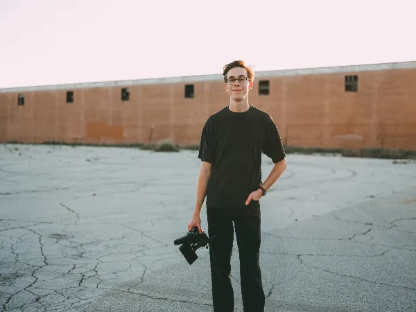 A young man wearing glasses and a black t-shirt stands in a paved lot holding a professional video camera.