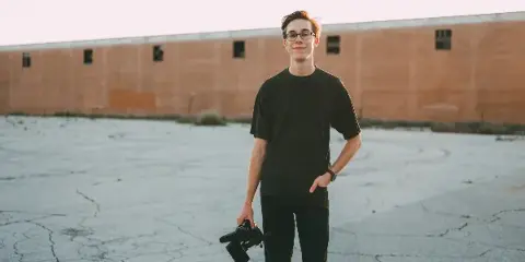 A young man wearing glasses and a black t-shirt stands in a paved lot holding a professional video camera.