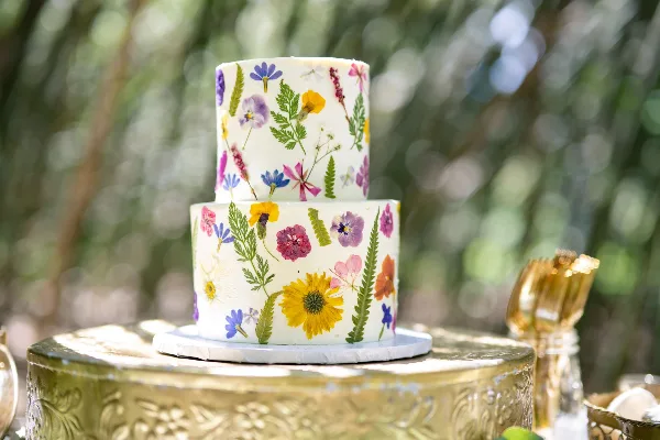 A two-tiered white wedding cake decorated with colorful pressed flowers and greenery, displayed on a gold stand.