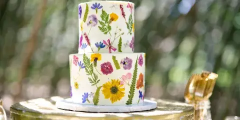 A two-tiered white wedding cake decorated with colorful pressed flowers and greenery, displayed on a gold stand.